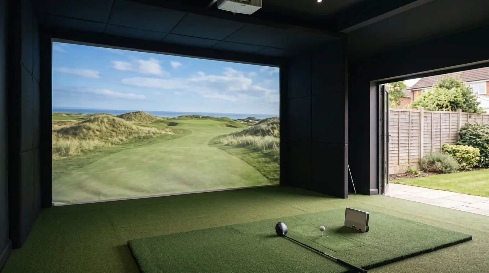 A home golf simulator in a garden room with the doors open to a British garden, the impact screen showing a links hole and a launch monitor beside a teed-up ball
